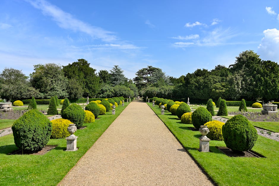 A tranquil lakeside garden scene during late afternoon, featuring a pebble and gravel shoreline in the foreground with a few weathered blue foam blocks and a wooden plank, possibly for garden or lands