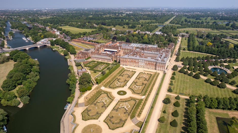 An aerial view of a large historic palace surrounded by meticulously maintained gardens, with symmetrical flower beds, neatly trimmed hedges, and pathways. The garden features a central circular featu