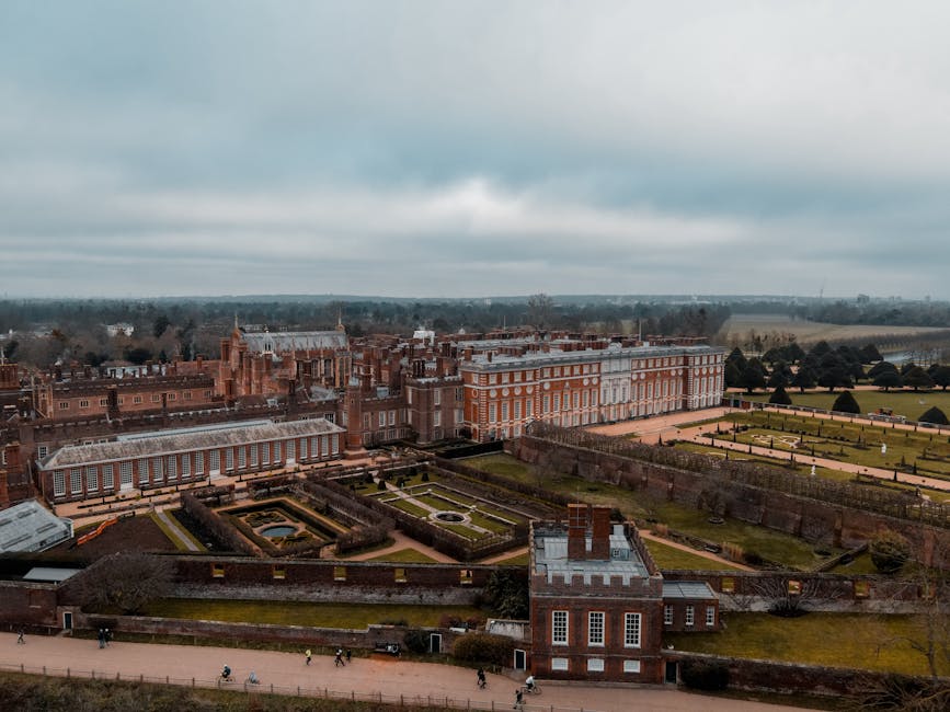 An aerial view of a large historic palace surrounded by meticulously maintained gardens, with symmetrical flower beds, neatly trimmed hedges, and pathways. The garden features a central circular featu